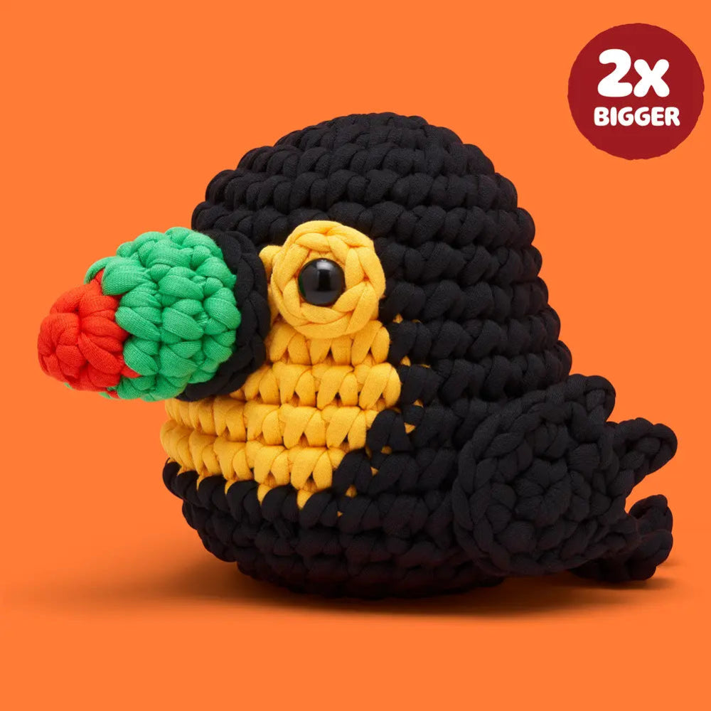 A crocheted plush toucan toy with a black body, yellow chest, green beak, and red beak tip, shown on an orange background with a red circular badge that reads '2x BIGGER'.