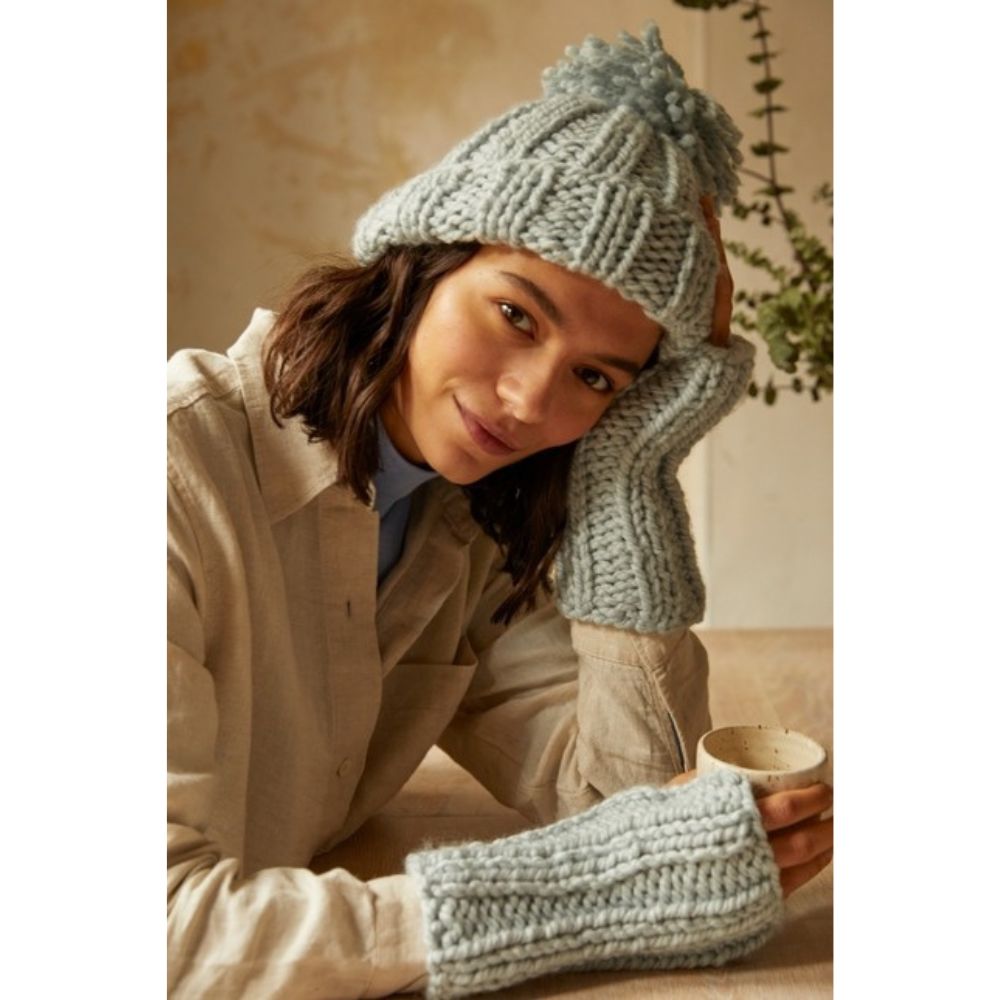 Woman wearing a cozy knit hat and mittens, holding a mug indoors.