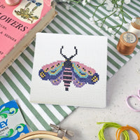 Cross-stitching project with a colorful moth design on a white fabric background on a table with a book, flowers and cross stitch supplies.