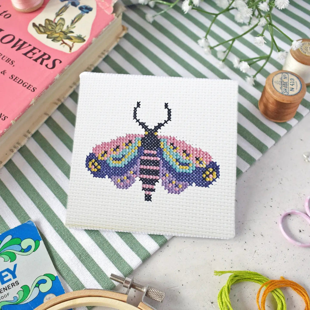 Cross-stitching project with a colorful moth design on a white fabric background on a table with a book, flowers and cross stitch supplies.