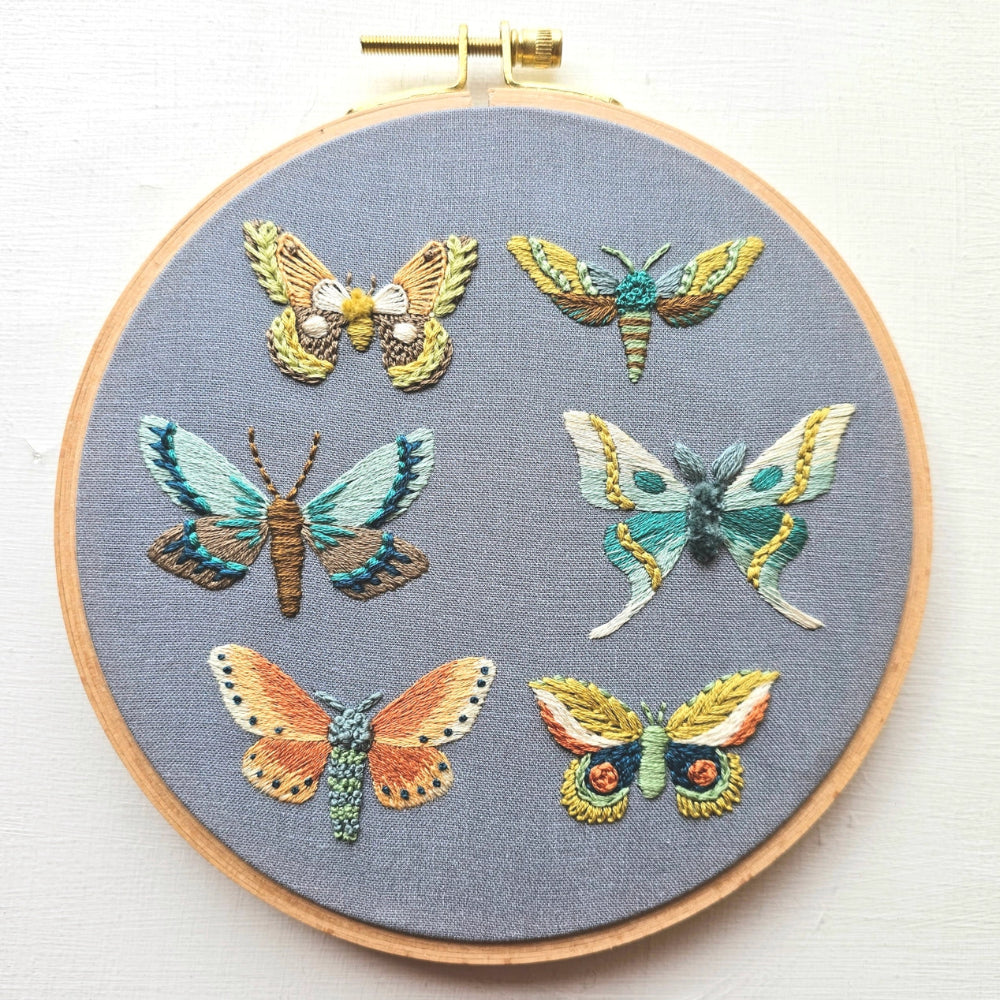 An embroidery hoop with six stitched moths in various colors against a grey background fabric
