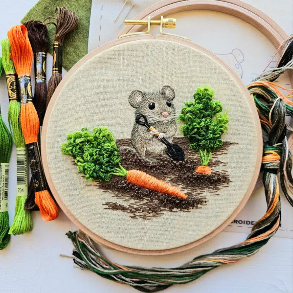 Embroidery kit of a mouse gardening and digging up carrots with strands of floss around it.