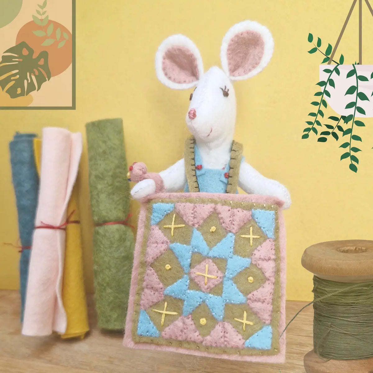 Handmade felt mouse holding a quilt with fabric rolls and a spool of thread in the background
