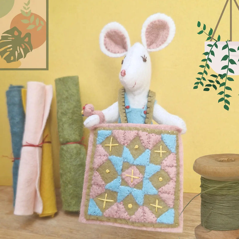 Handmade felt mouse holding a quilt with fabric rolls and a spool of thread in the background