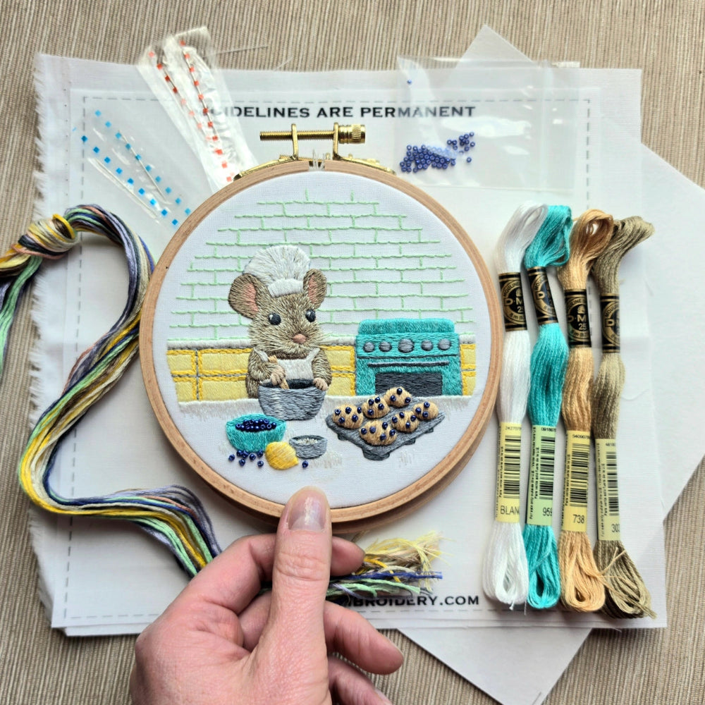 An embroidery hoop containing fabric with a stitched mouse baking muffins in a cute kitchen