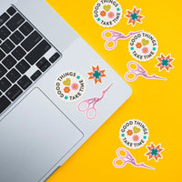 Several stickers, including  stork scissors, a quilting star and the phrase "Good Things Take Time" are placed on a laptop and against a yellow background. 