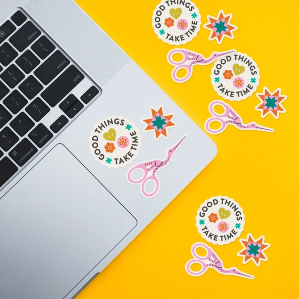 Several stickers, including  stork scissors, a quilting star and the phrase "Good Things Take Time" are placed on a laptop and against a yellow background. 