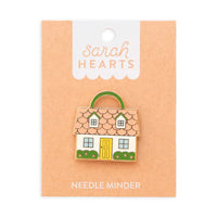 A needle minder shaped like a house with a  pink roof, yellow door, and green shrubs on brown Sarah Hearts packaging. 