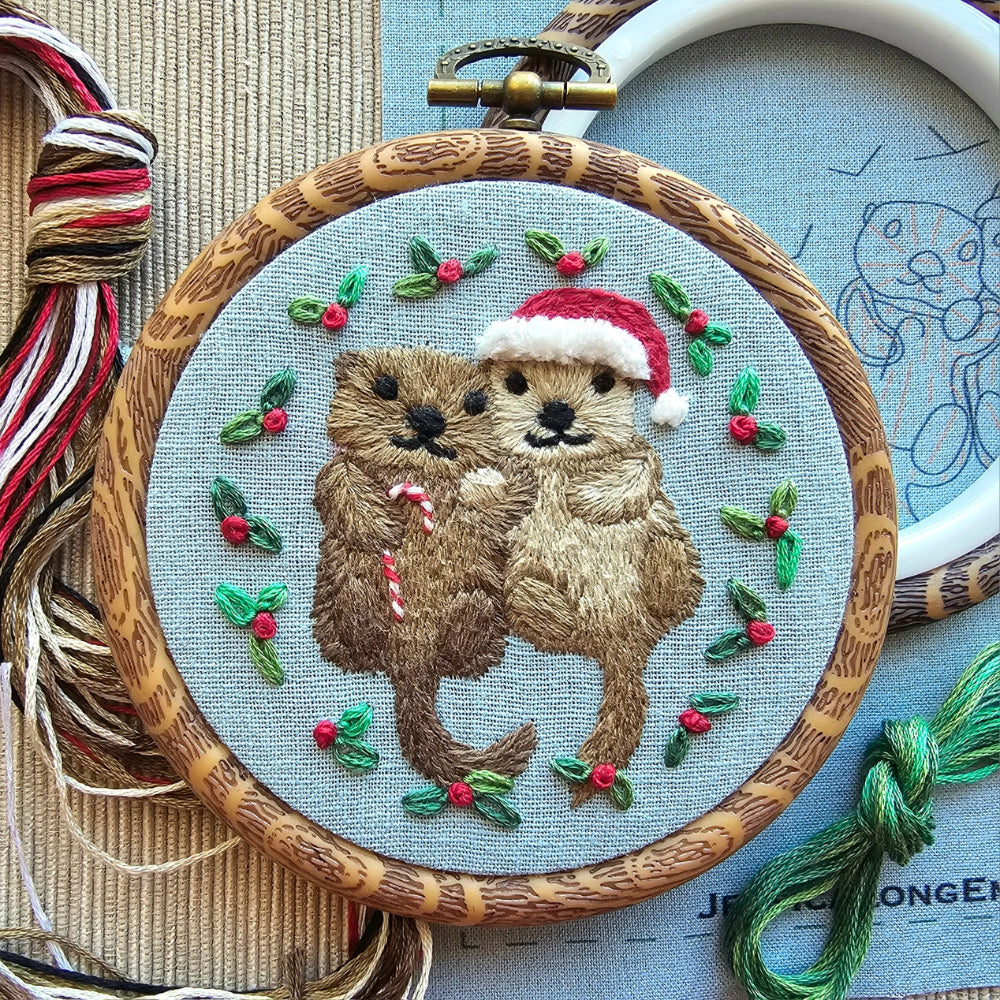 A small embroidery hoop holding pale blue fabric with an embroidered pair of otters, one wearing a santa hat and one carrying a candy cane