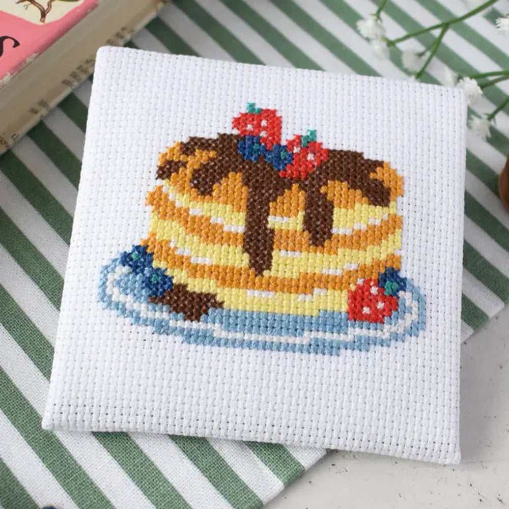 Cross-stitched design of a pancake with syrup and berries on a white fabric background