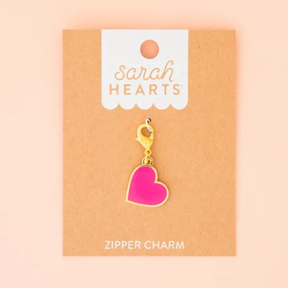A pink heart charms with a golden border and clasp is on brown Sarah Hearts packaging on a light pink background