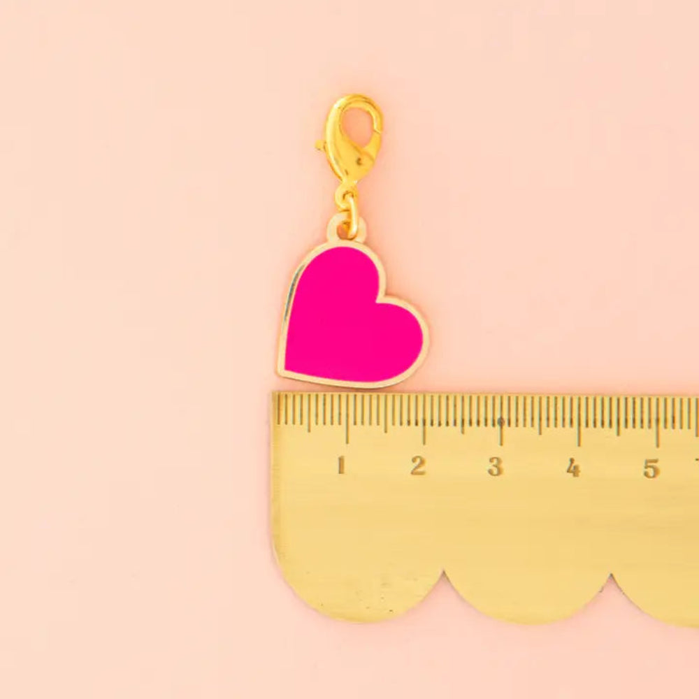 A pink heart charm with a golden clasp is on top of a scalloped edge ruler against a light pink background.