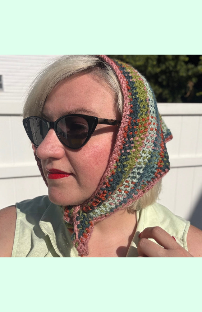Blonde woman with black sunglasses and red lips wearing a crocheted multicolored plaid bandana tied around her chin.