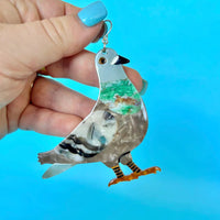 Flat pigeon-shaped acetate earring with silver ear hook, held in a hand with blue nail polish.