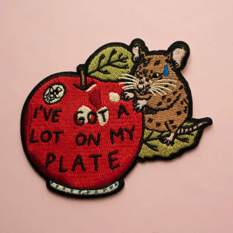 Woven patch depicting a cute little mouse trying to eat a big apple. The apple has text on it that says 'I've Got A Lot On My Plate'.