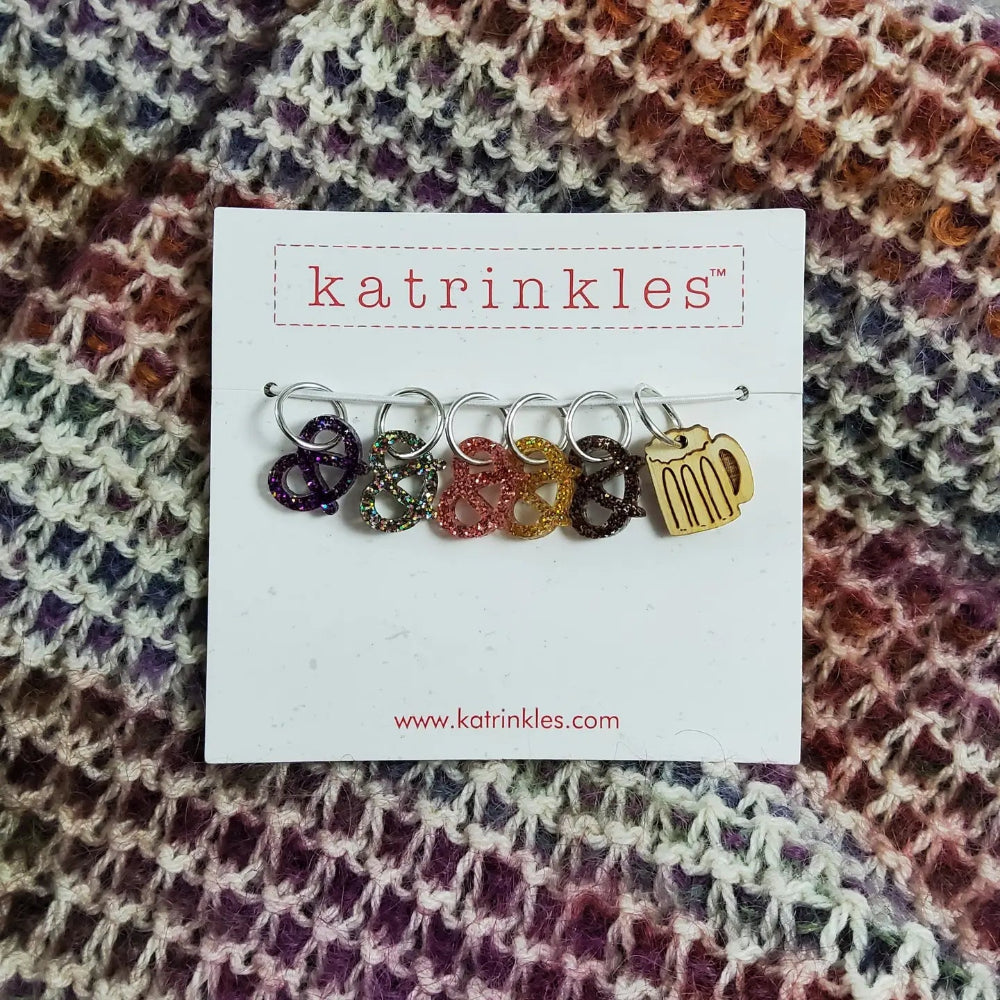 Set of colorful pretzel and beer shaped stitch markers on a card with 'katrinkles' branding, placed on a fabric background.