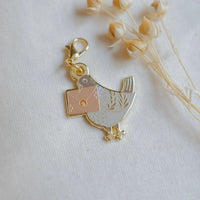 A small gold-tone bird charm carrying a square envelope, photographed on a light fabric surface with dried flowers in the background.
