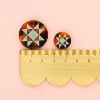 A tortoise shell button with mint green geometric star pattern is next to a smaller sized button on top of a scalloped edge ruler on a pink background.