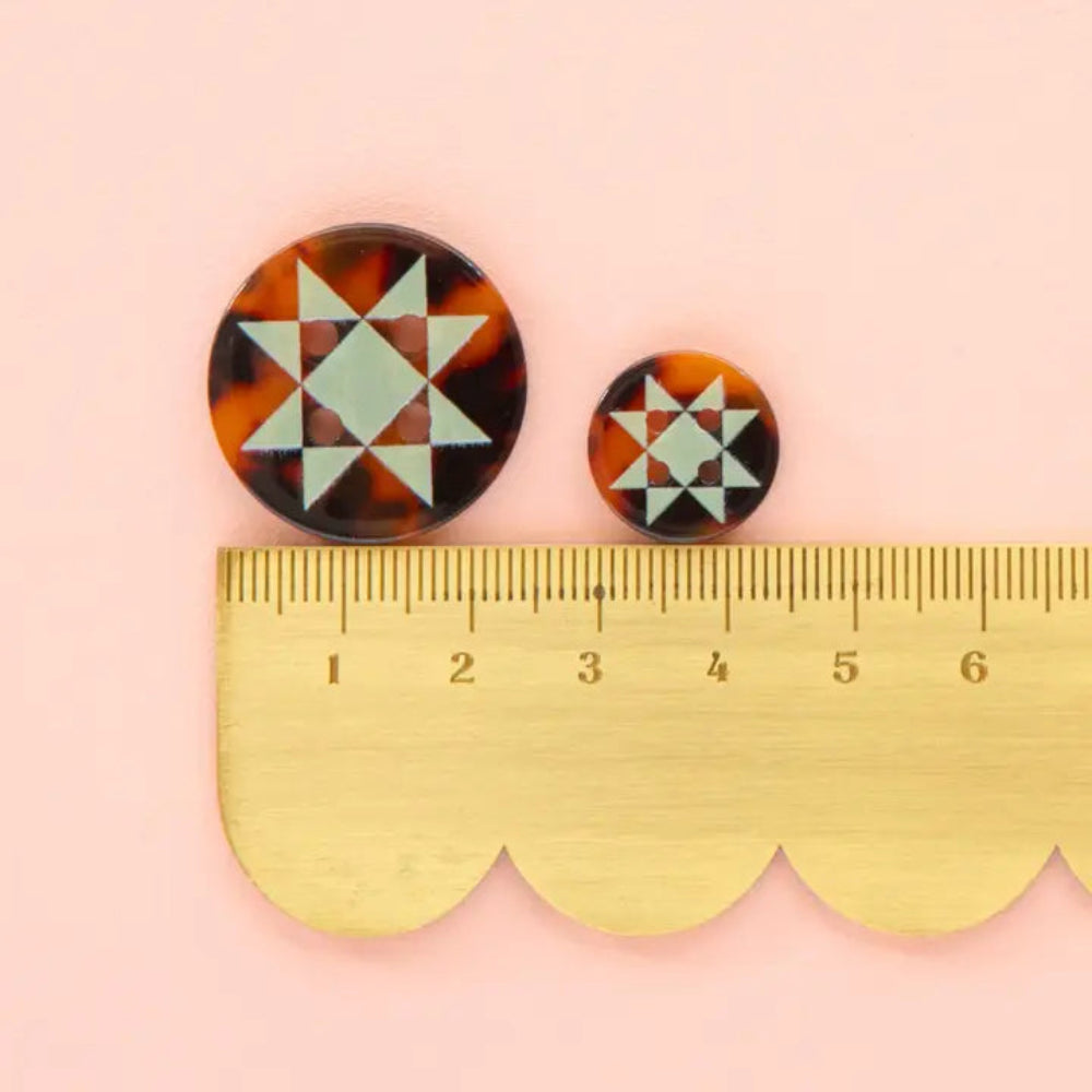 A tortoise shell button with mint green geometric star pattern is next to a smaller sized button on top of a scalloped edge ruler on a pink background.