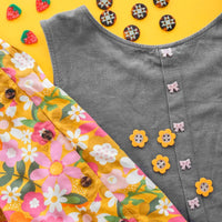 A denim dress with pink bows is laid next a bright floral fabric featuring yellow, pink, and white flowers and tortoise shell buttons. Strawberry buttons and star-patterned buttons decorate the yellow background.