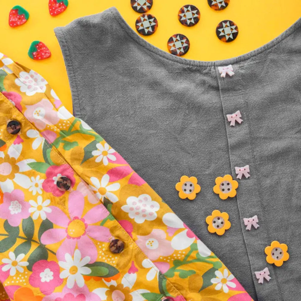 A denim dress with pink bows is laid next a bright floral fabric featuring yellow, pink, and white flowers and tortoise shell buttons. Strawberry buttons and star-patterned buttons decorate the yellow background.
