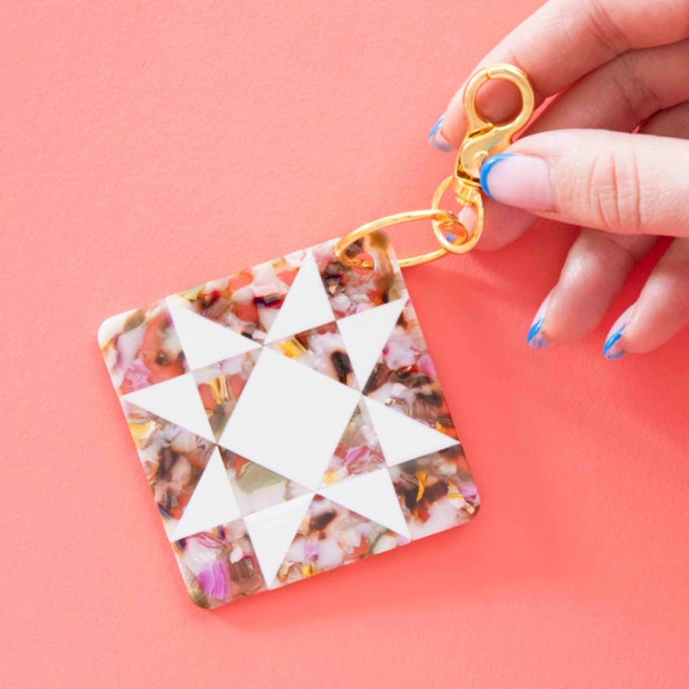 A hand with blue-tipped nails holds a multicolor  keychain with a quilting star pattern in white against a pink background. 