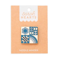Square needle minder with different designs: blue flower, a quilting star, triangles, and a rainbow on brown backing labeled "Sarah Hearts"