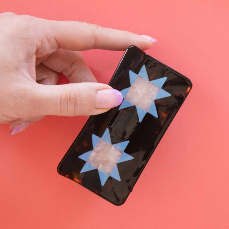 A hand with pink-tipped nails holds a tortoise hair clip adorned with two blue quilt star shapes, against a coral background. 