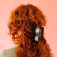 A person with red wavy hair wears a  tortoise hair clip adorned with two blue quilt star shapes, against a coral background. 