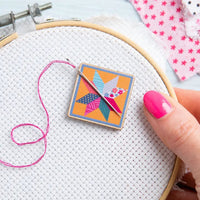 Embroidery hoop with cross-stitching fabric, colorful patchwork square magnet, and pink thread.