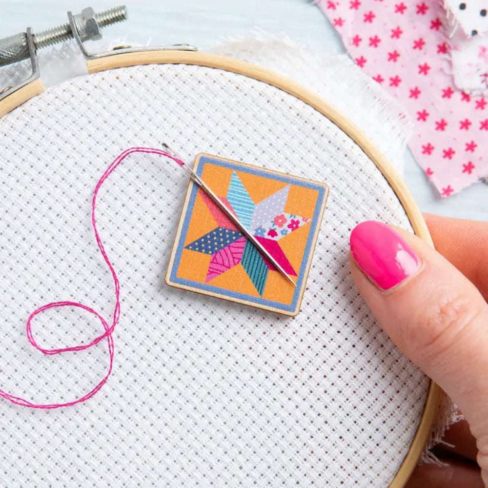 Embroidery hoop with cross-stitching fabric, colorful patchwork square magnet, and pink thread.