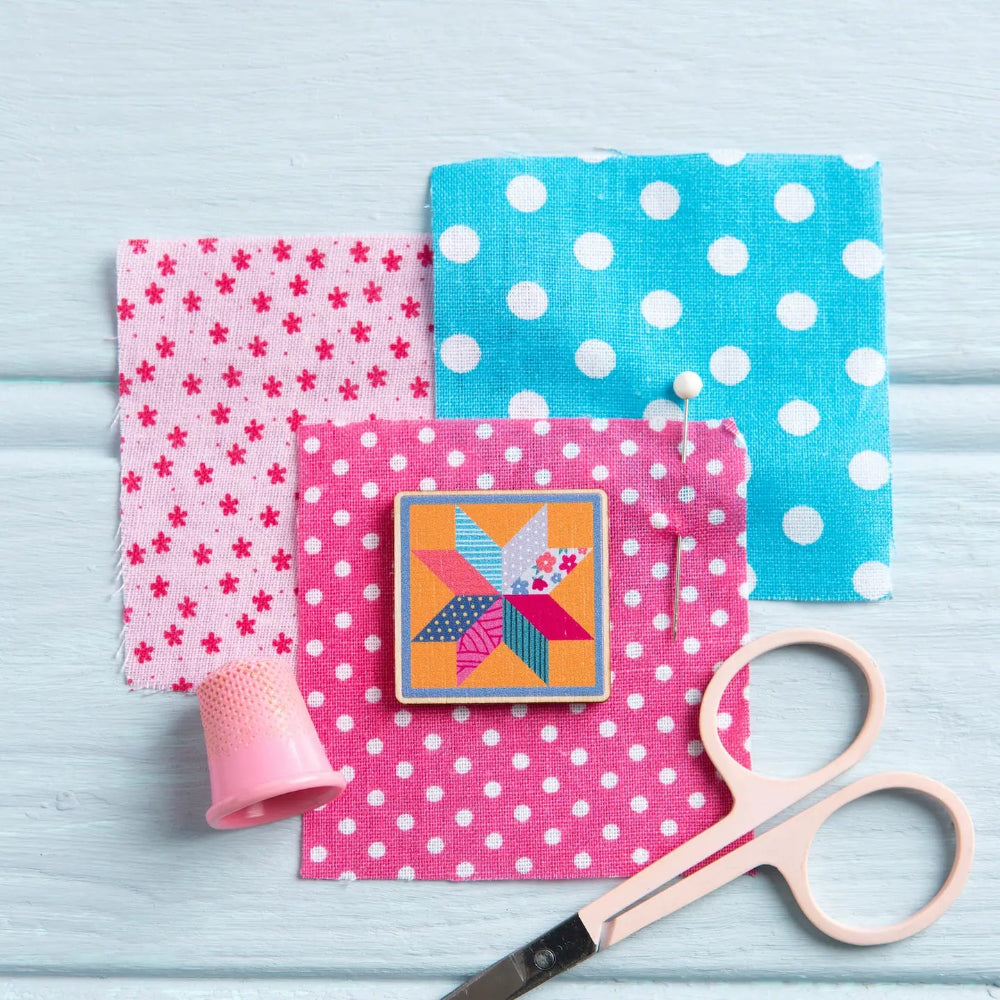 Colorful fabric squares with patterns, a quilt magnetic pin, and scissors on a light blue background