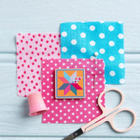 Colorful fabric squares with patterns, a quilt magnetic pin, and scissors on a light blue background