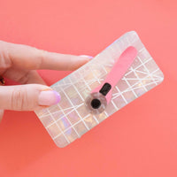 A hand holds a holographic cutting mat claw clip with a pink rotary cutter attached, set against a vibrant coral background. 