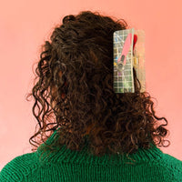 A person with brown curly hair and a green sweater wears a holographic claw clip with a pink rotary cutter design, set against a vibrant coral background. 