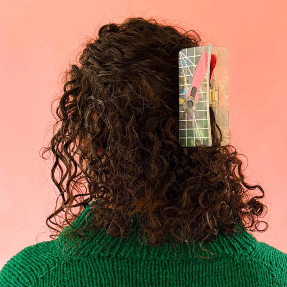 A person with brown curly hair and a green sweater wears a holographic claw clip with a pink rotary cutter design, set against a vibrant coral background. 