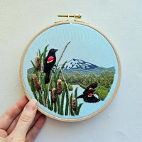 Embroidery hoop with a scenic design of birds, mountains, and plants held by a hand on a light background.