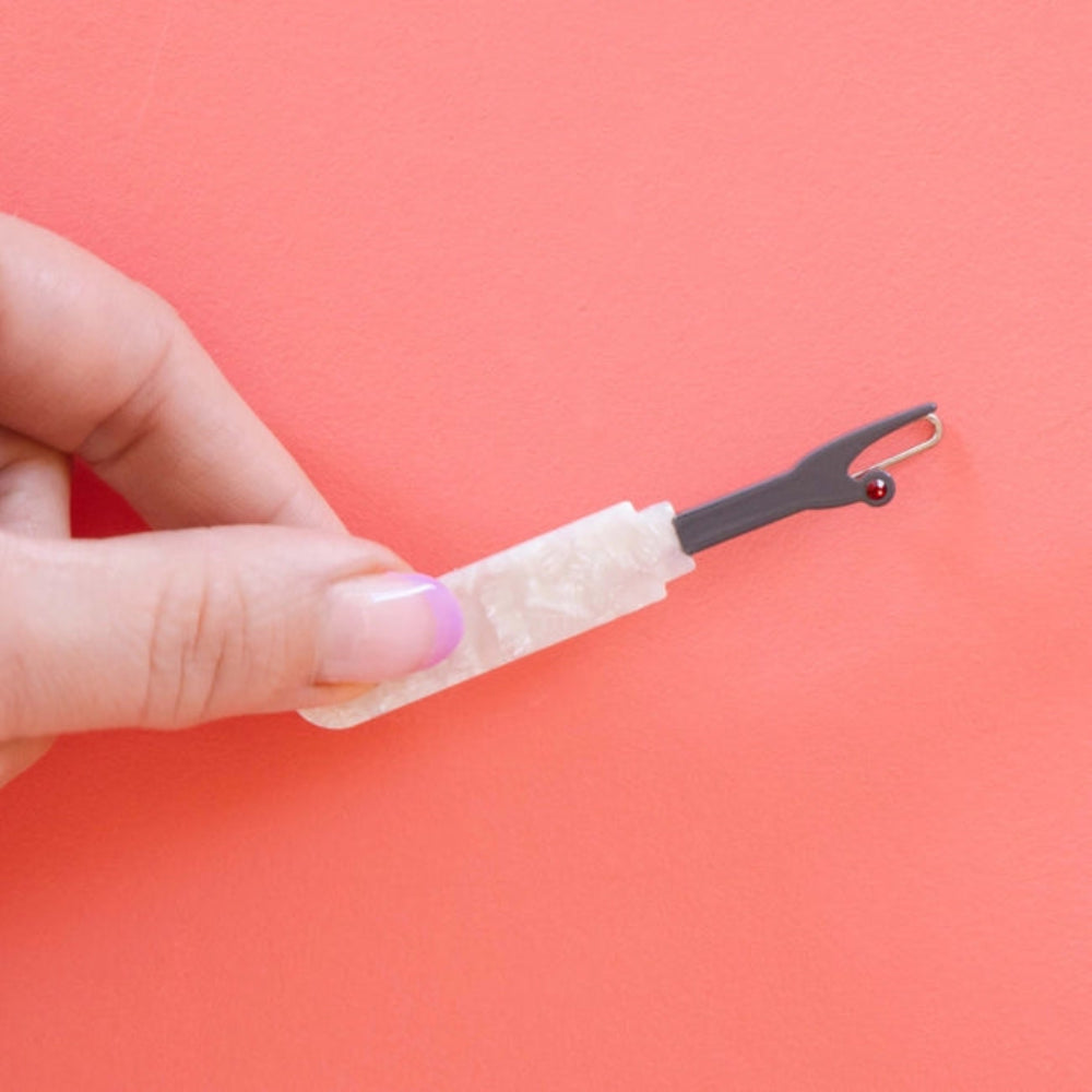 A hand holds a hair clip shaped like a seam ripper with a pearlized handle  and red gem point. 