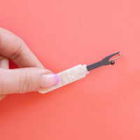 A hand holds a hair clip shaped like a seam ripper with a pearlized handle  and red gem point. 