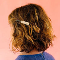 A person with short, wavy, brown hair wears a hair clip shaped like a seam ripper with a pearlized handle against a pink brackground. 