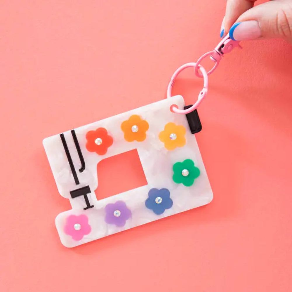 Pearlized sewing machine keychain with colorful daisy decorations held by a hand against a coral background.