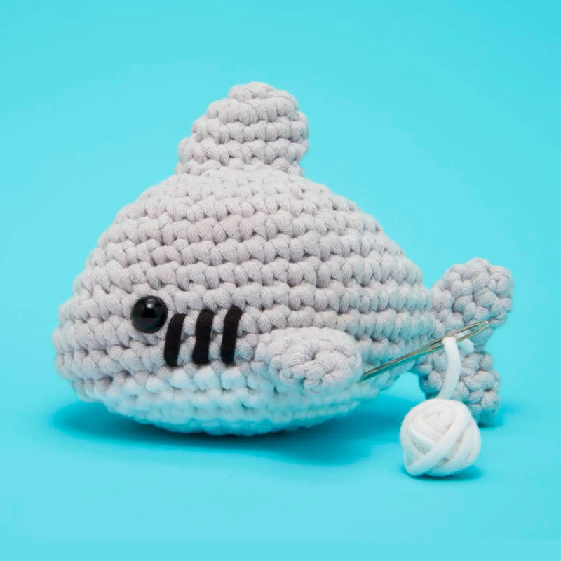 A crocheted plush shark toy in light gray yarn with black stitched details, shown on a bright cyan background with a yarn ball and needle visible.