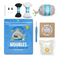 The Woobles Learn to Crochet Kit for Beginners, featuring a blue pouch with a crocheted shark photo, black and white yarn, a gray yarn ball, a blue crochet hook, a small booklet, and other accessories on a white background.