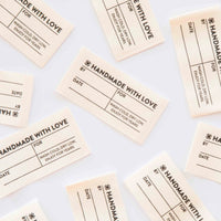 Scattered cream-colored fabric labels, titled "Handmade with Love" with spaces for "By," "Date," and "For." Instructions: "Wash cold, dry low. Enjoy For Years"