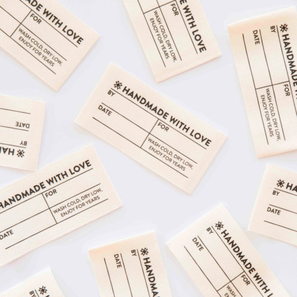 Scattered cream-colored fabric labels, titled "Handmade with Love" with spaces for "By," "Date," and "For." Instructions: "Wash cold, dry low. Enjoy For Years"