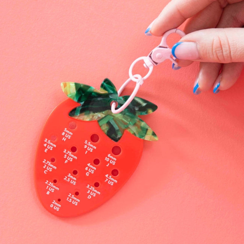 A hand  holds a strawberry-shaped tool for measuring hook sizes with labeled holes in white text. 