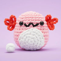 A pink crocheted axolotl plush toy with red ear, black eyes and smile, shown on a light purple background with a small white yarn ball beside it.