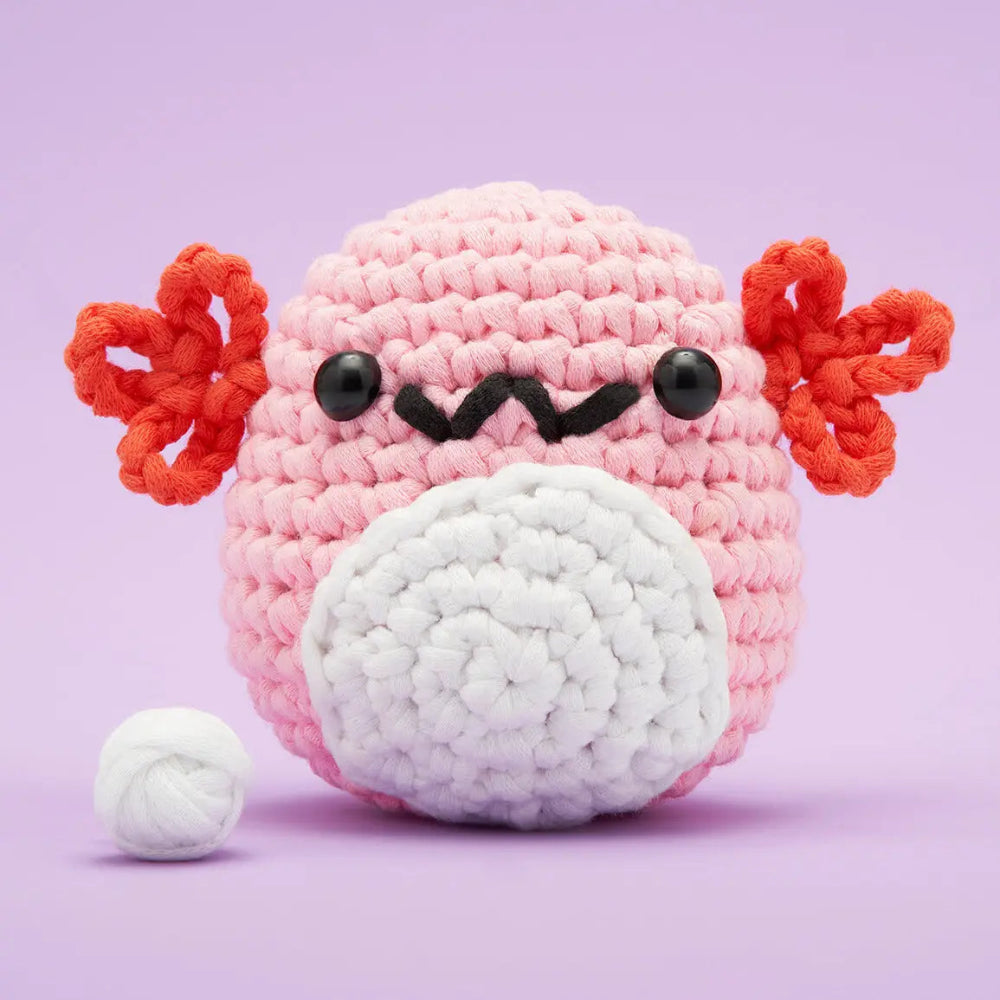 A pink crocheted axolotl plush toy with red ear, black eyes and smile, shown on a light purple background with a small white yarn ball beside it.