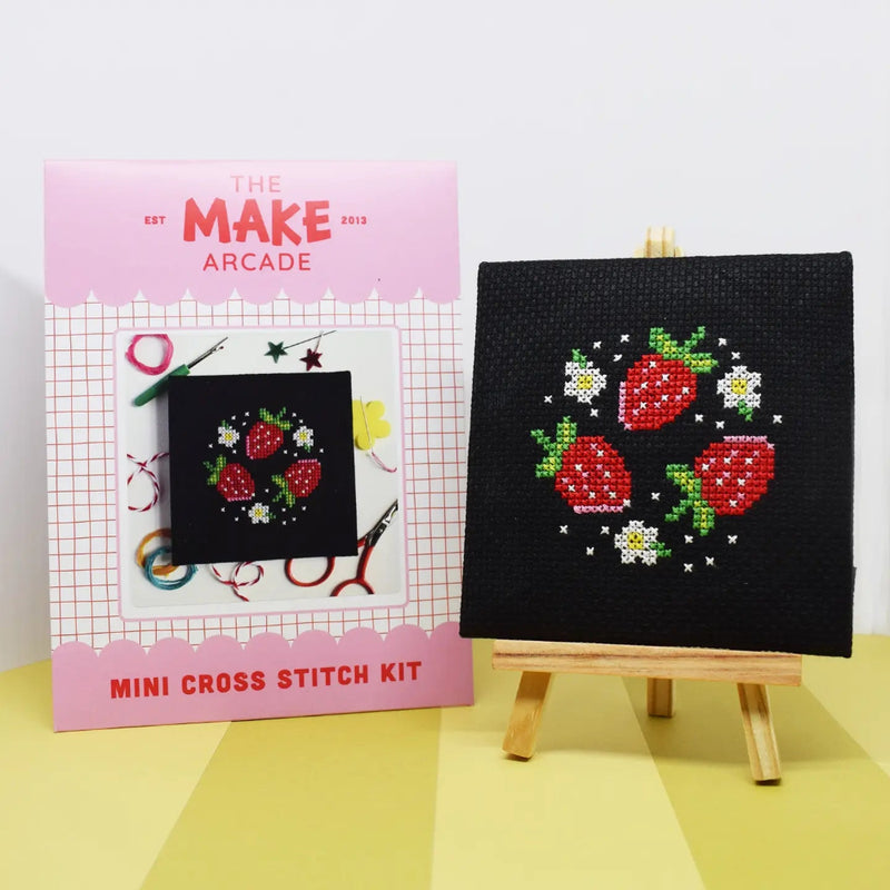 Mini cross stitch kit with strawberry design on black fabric, next to a cross stitch kit with Make Arcade packaging. 