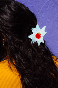 Person with a star-shaped hair clip featuring a red tomato design against a purple textured background